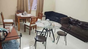 A living room with a round glass table, wooden chairs, a brown leather sofa, and a dining table with chairs.
