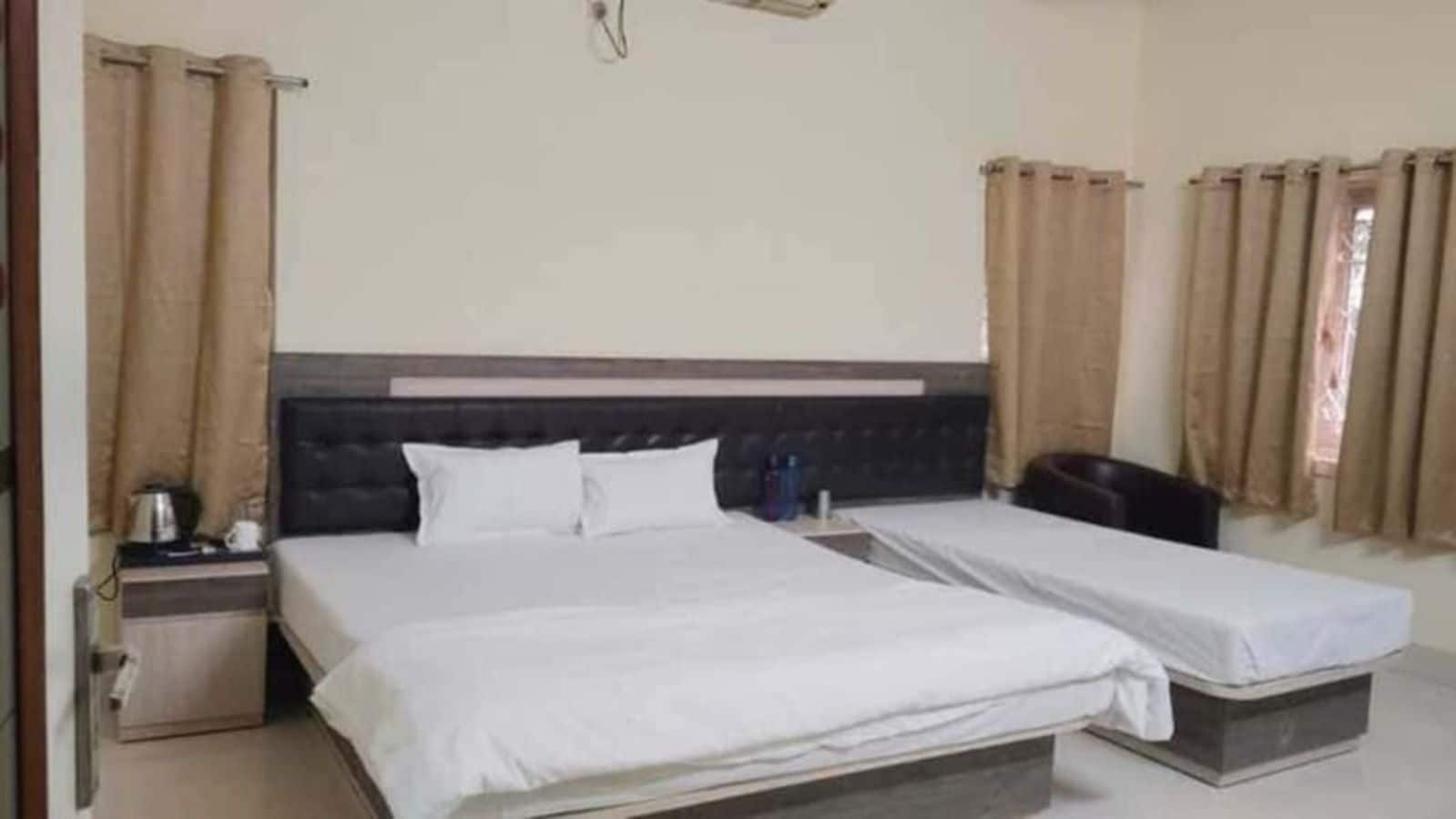 Ein Hotelzimmer mit einem großen Bett, einem kleineren Bett, einem Nachttisch, einer Lampe und Vorhängen.