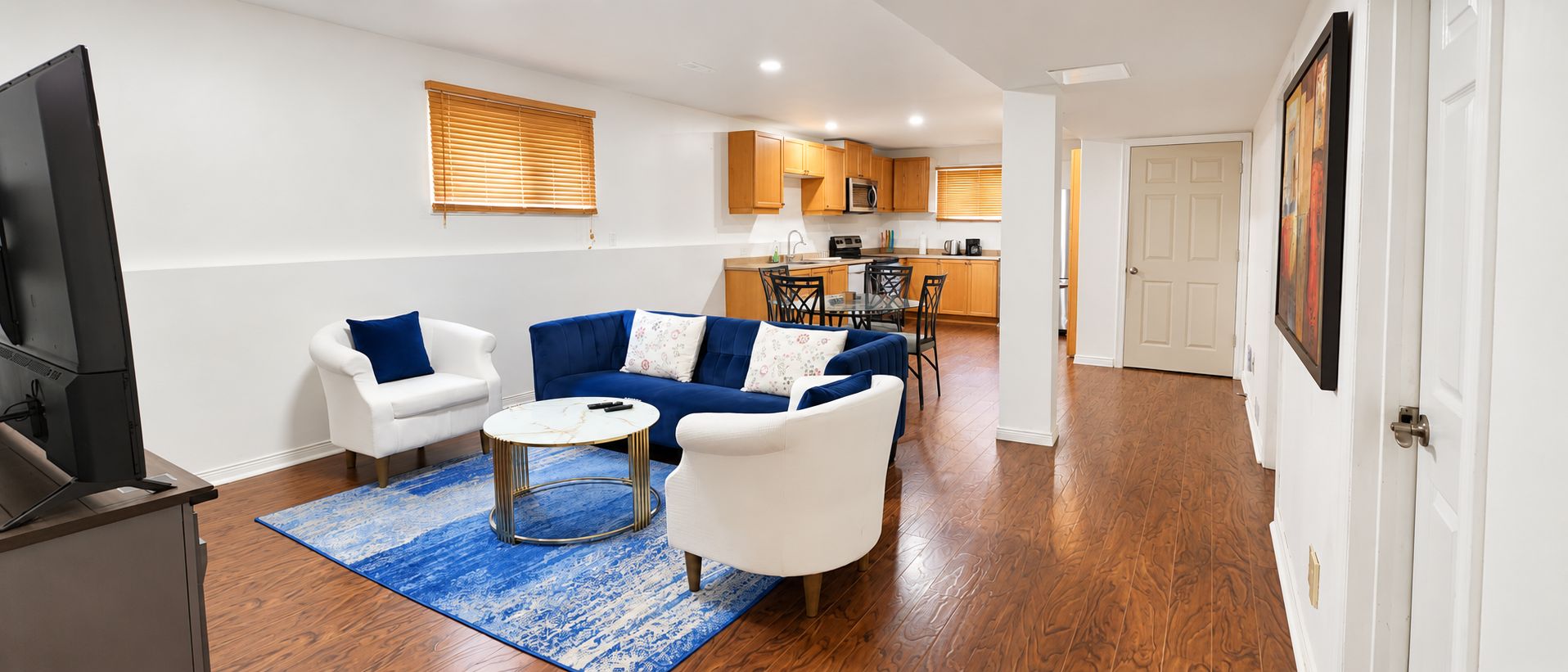 A living room with a sofa, armchair, and coffee table, a kitchen in the background, and a door leading to another room.