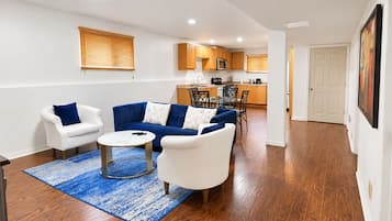 A living room with a sofa, armchair, and coffee table, a kitchen in the background, and a door leading to another room.