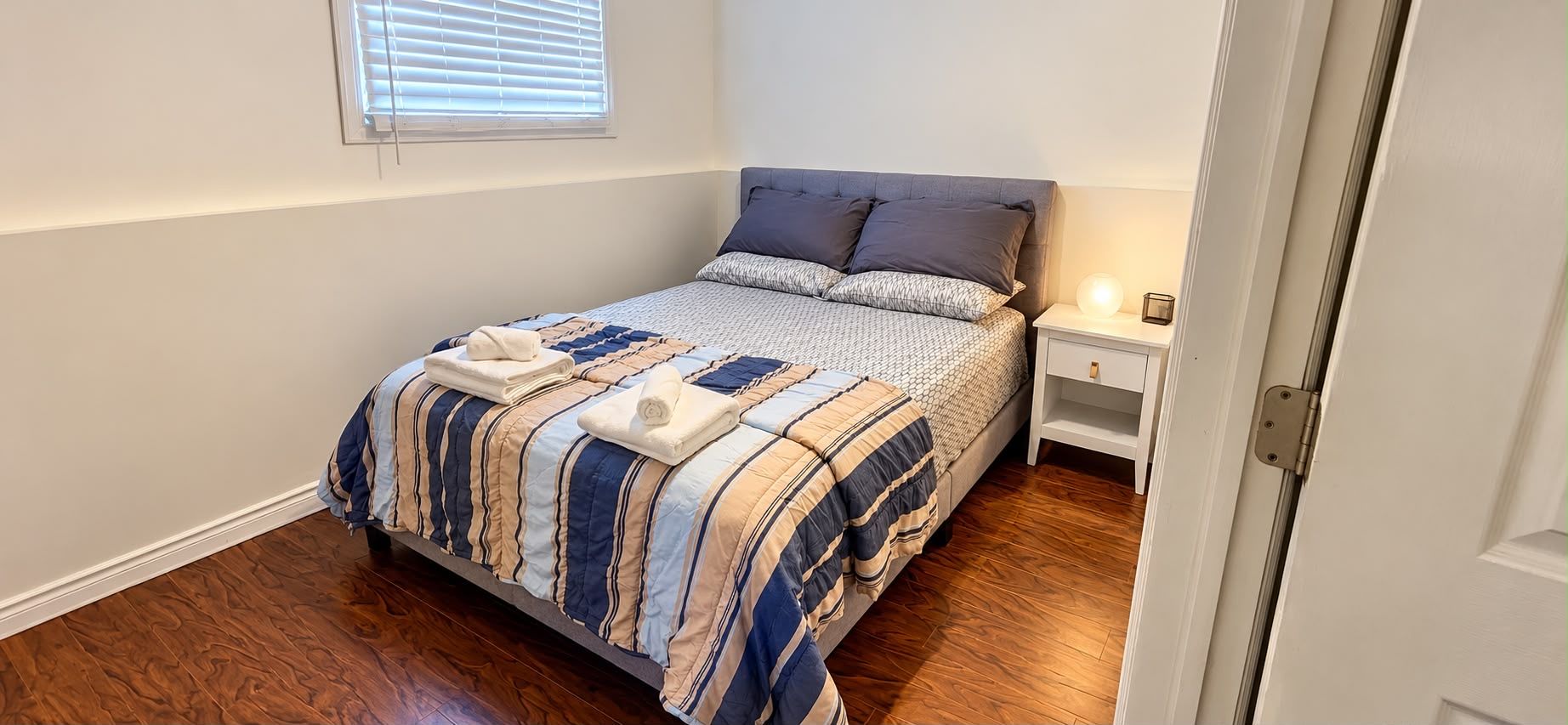 A bedroom with a bed, a nightstand, a window with blinds, and a door.