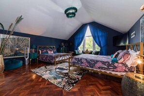 A bedroom with two beds, a large window with blue curtains, a peacock painting, and a herringbone floor.