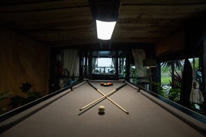 A pool table with a cue ball and two cues, surrounded by plants and a window with a view of greenery.