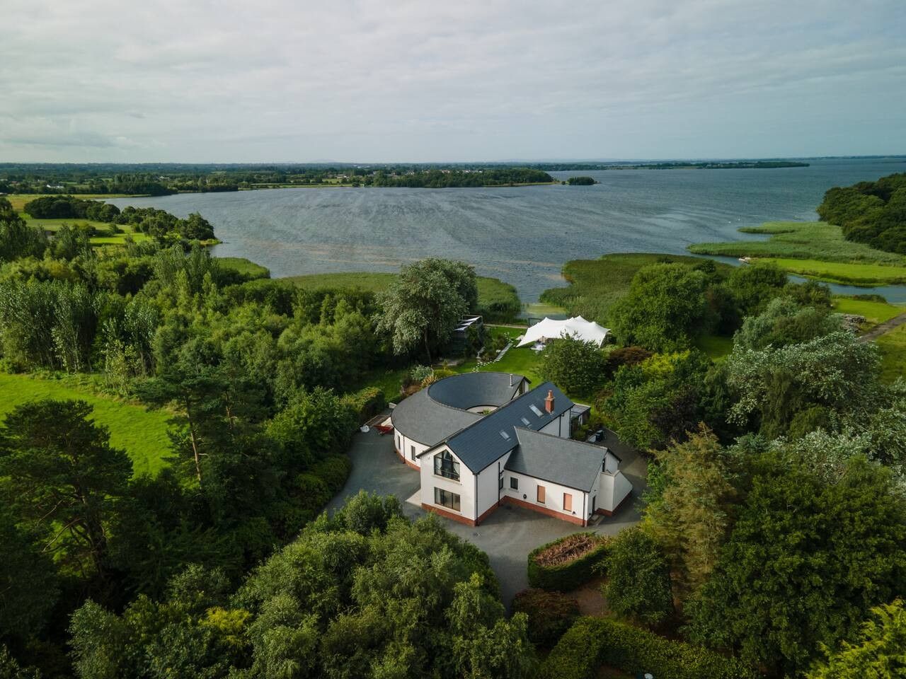 A large house with a spacious driveway, surrounded by lush greenery and a body of water in the background.