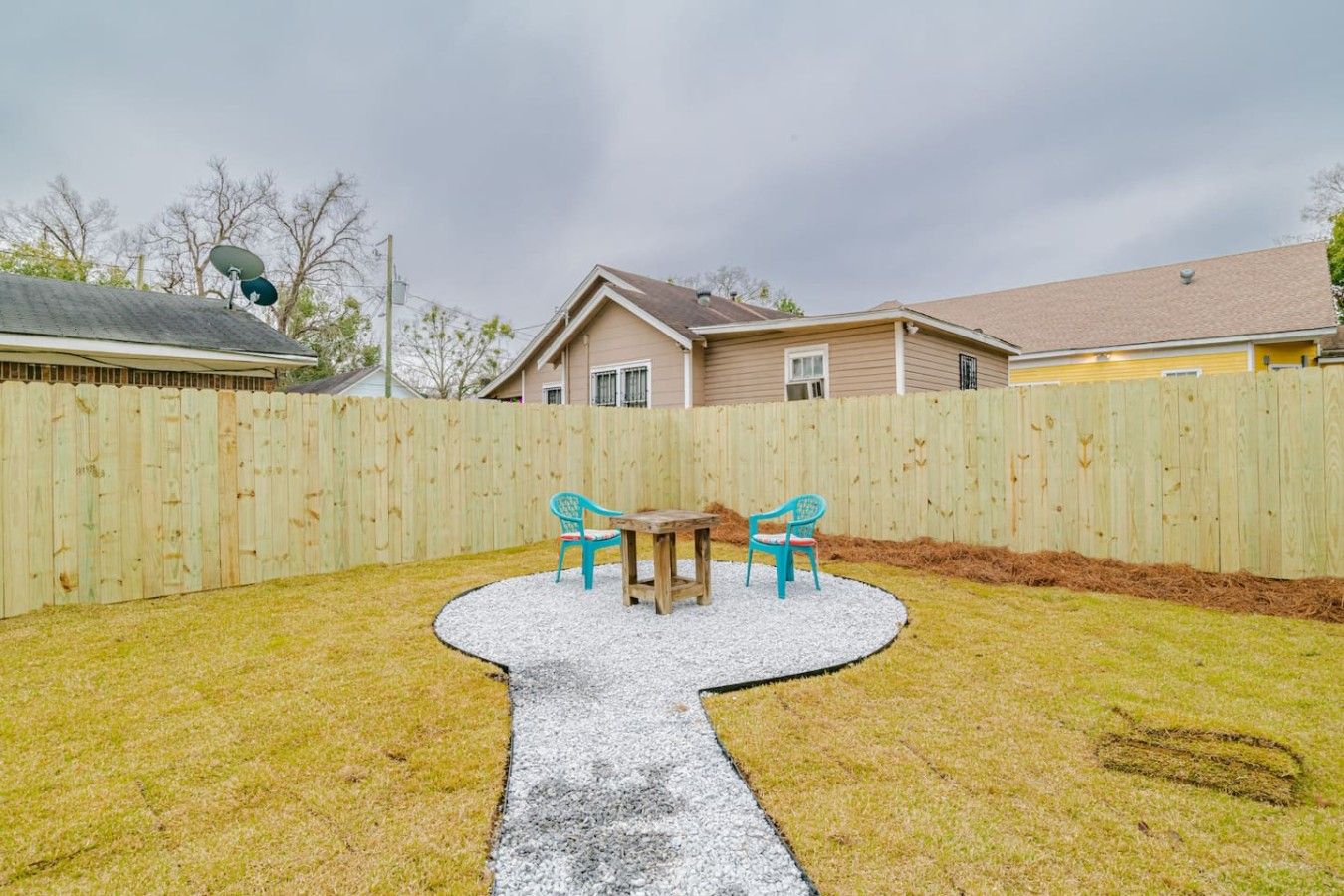 Un patio trasero con una mesa y sillas de madera, un sendero de grava y una cerca de madera.