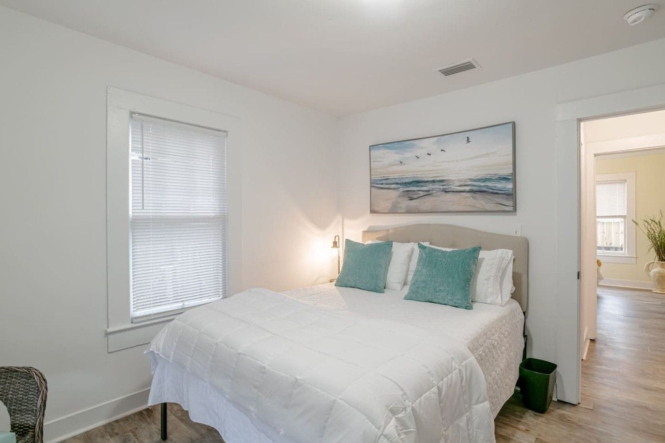 A bedroom with a bed, a window, a painting, and a door leading to another room.