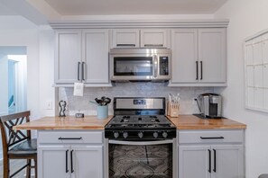 Una cocina con gabinetes blancos, un microondas de acero inoxidable, una estufa a gas y una cafetera.