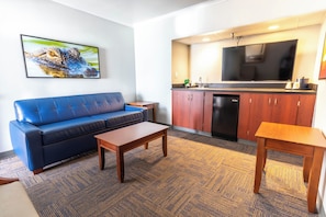 A living room with a blue sofa, a wooden coffee table, and a kitchen area with a large TV and cabinets.