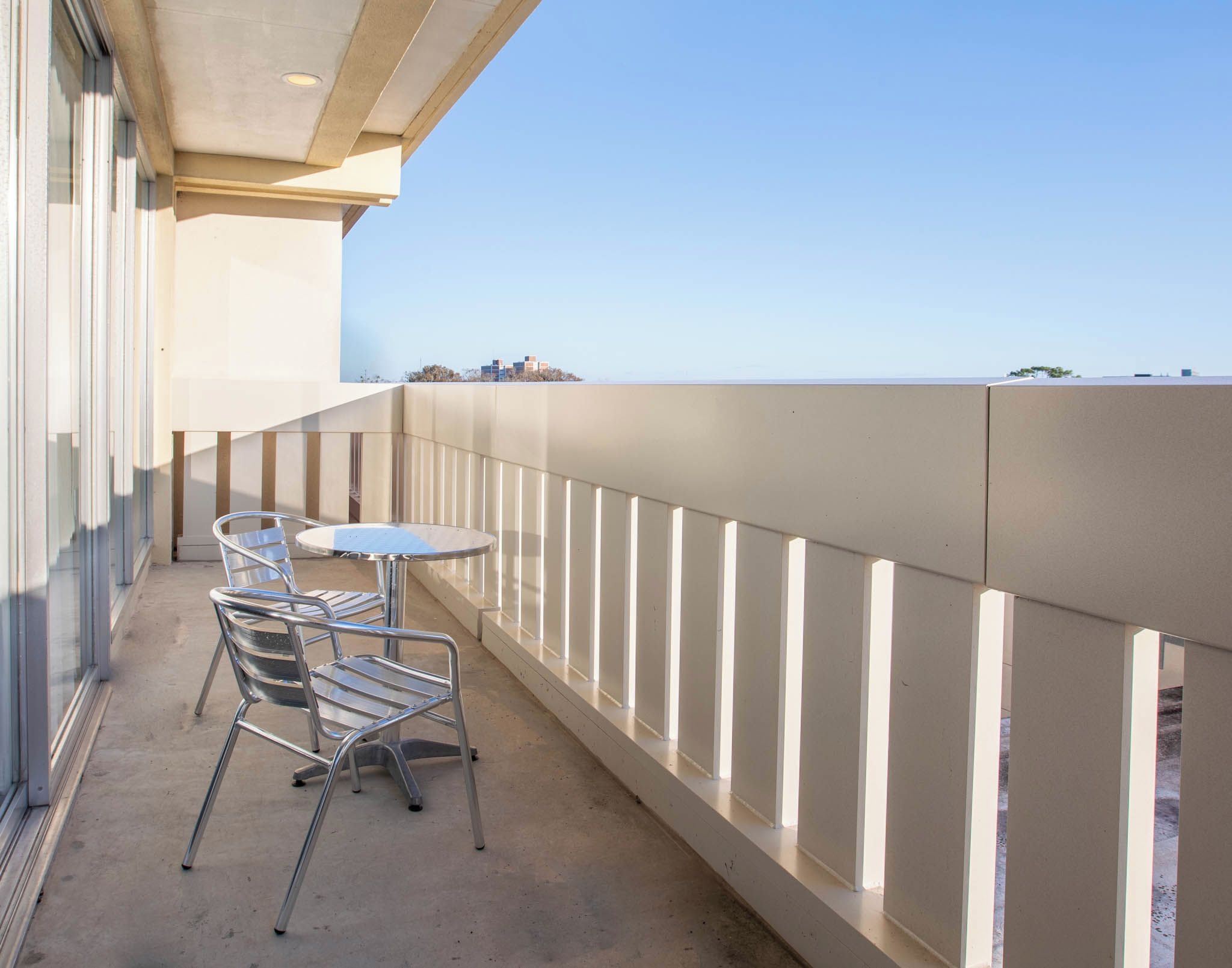 Un balcón con mesa y sillas, con vistas a un cielo despejado y edificios lejanos.