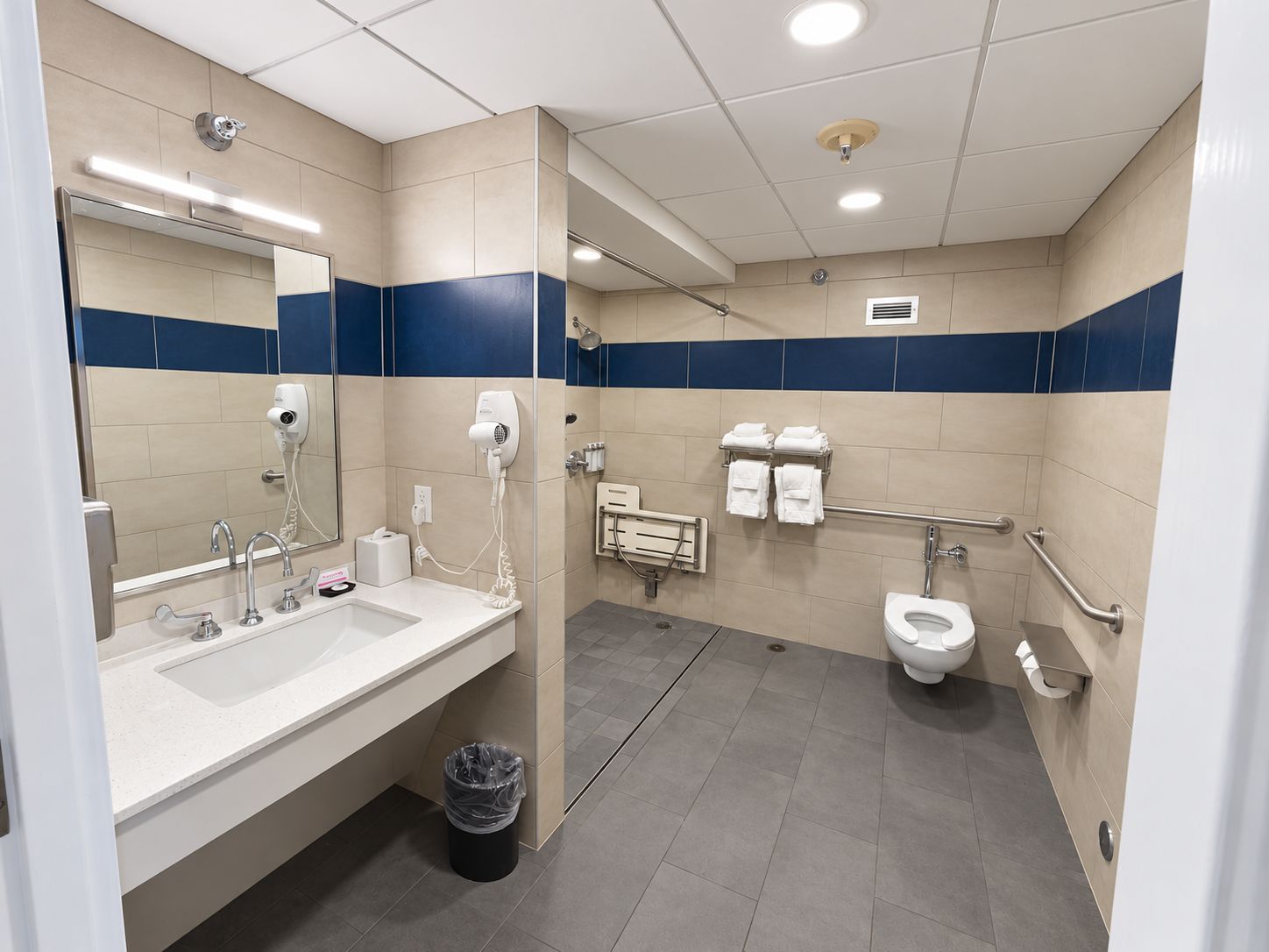 A modern bathroom with a dual sink vanity, a mirror, a hair dryer, a toilet, and a trash bin.