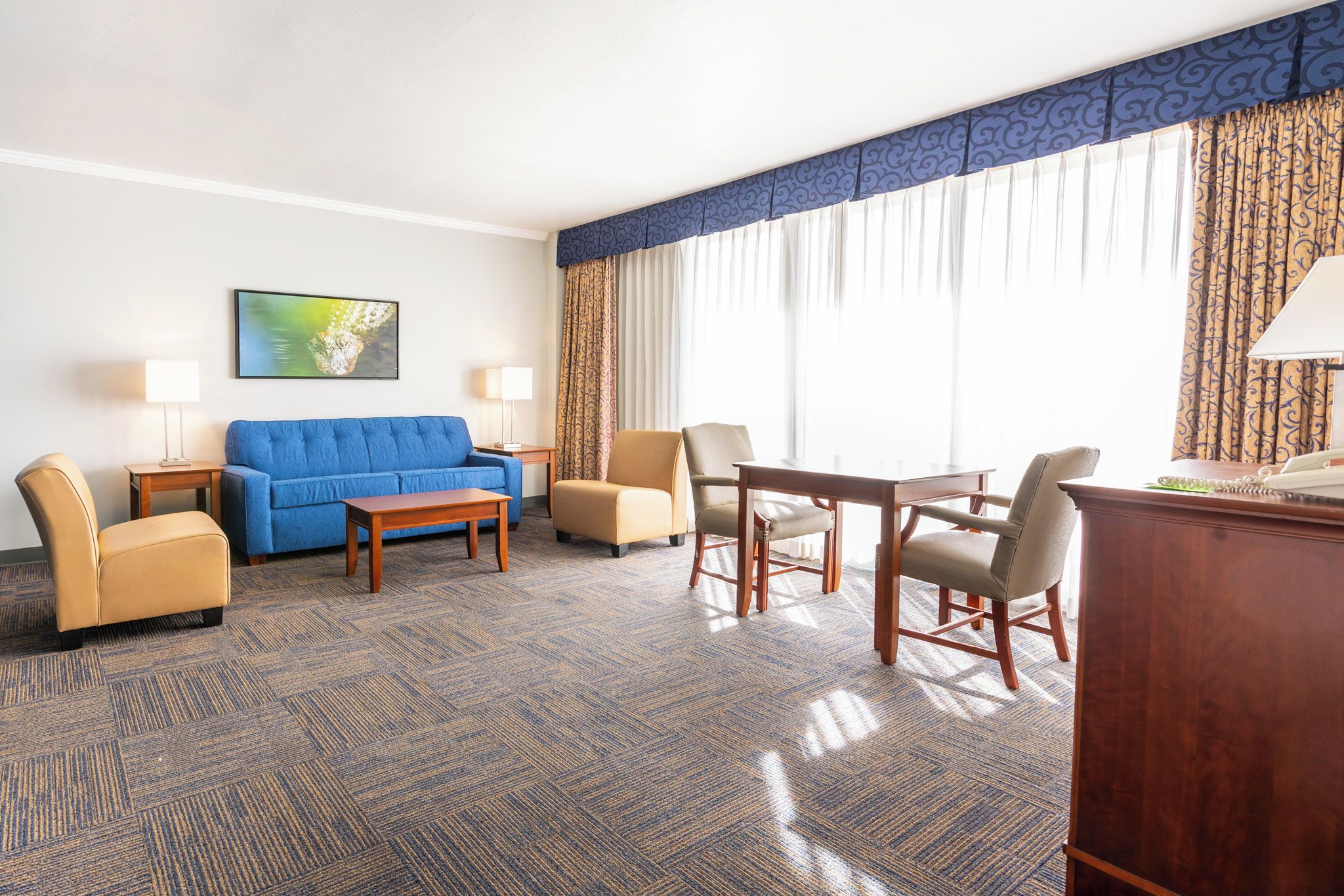 Habitación de hotel con un sofá azul, dos sillones, una mesa de centro de madera, un escritorio con teléfono y un ventanal con cortinas.