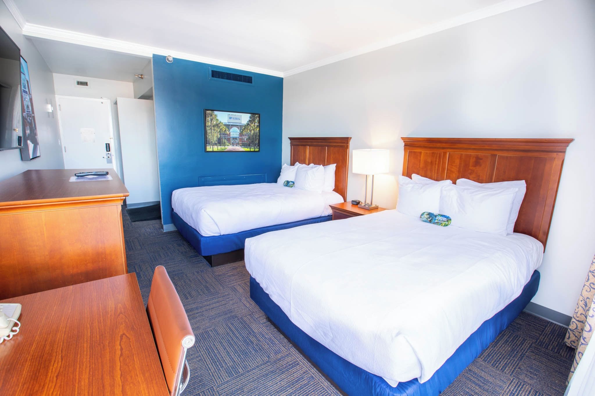 Habitación de hotel con dos camas, un escritorio, una silla, una lámpara y un cuadro en la pared.