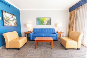 Habitación de hotel con un sofá azul, dos sillones, una mesa de centro de madera y dos mesitas de noche con lámparas.