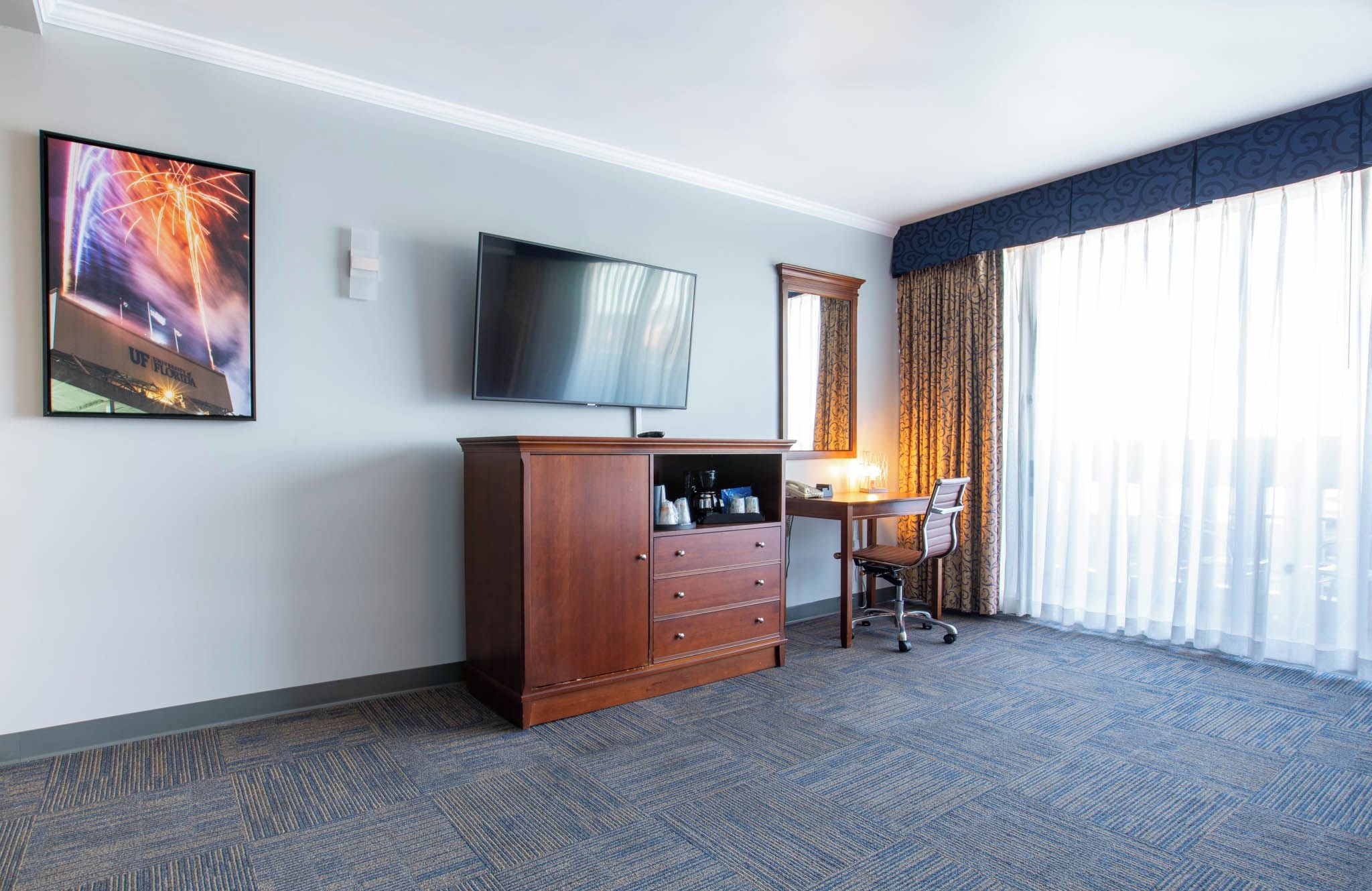 Habitación de hotel con televisión de pantalla plana, cómoda de madera, escritorio con silla y ventana grande con cortinas.