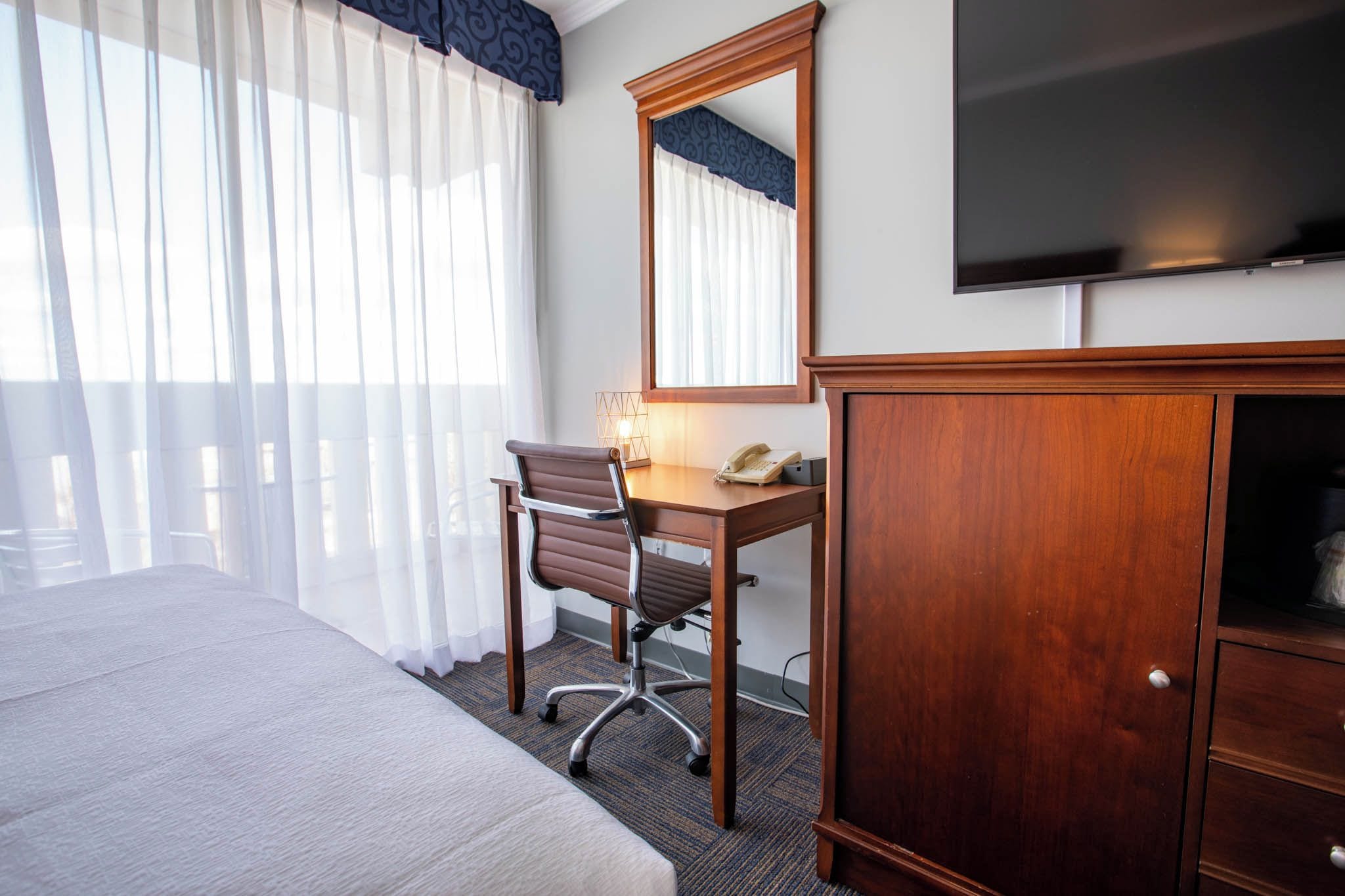 A hotel room with a bed, a desk with a chair, a telephone, a mirror, and a television on the wall.