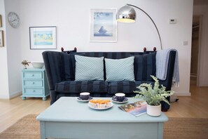 Uma sala de estar com um sofá, uma mesa de centro com doces, um relógio e quadros nas paredes.