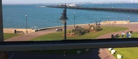 A view from a window overlooking a coastal area with a pier extending into the water, a grassy area with a lamp post, and a clear sky.
