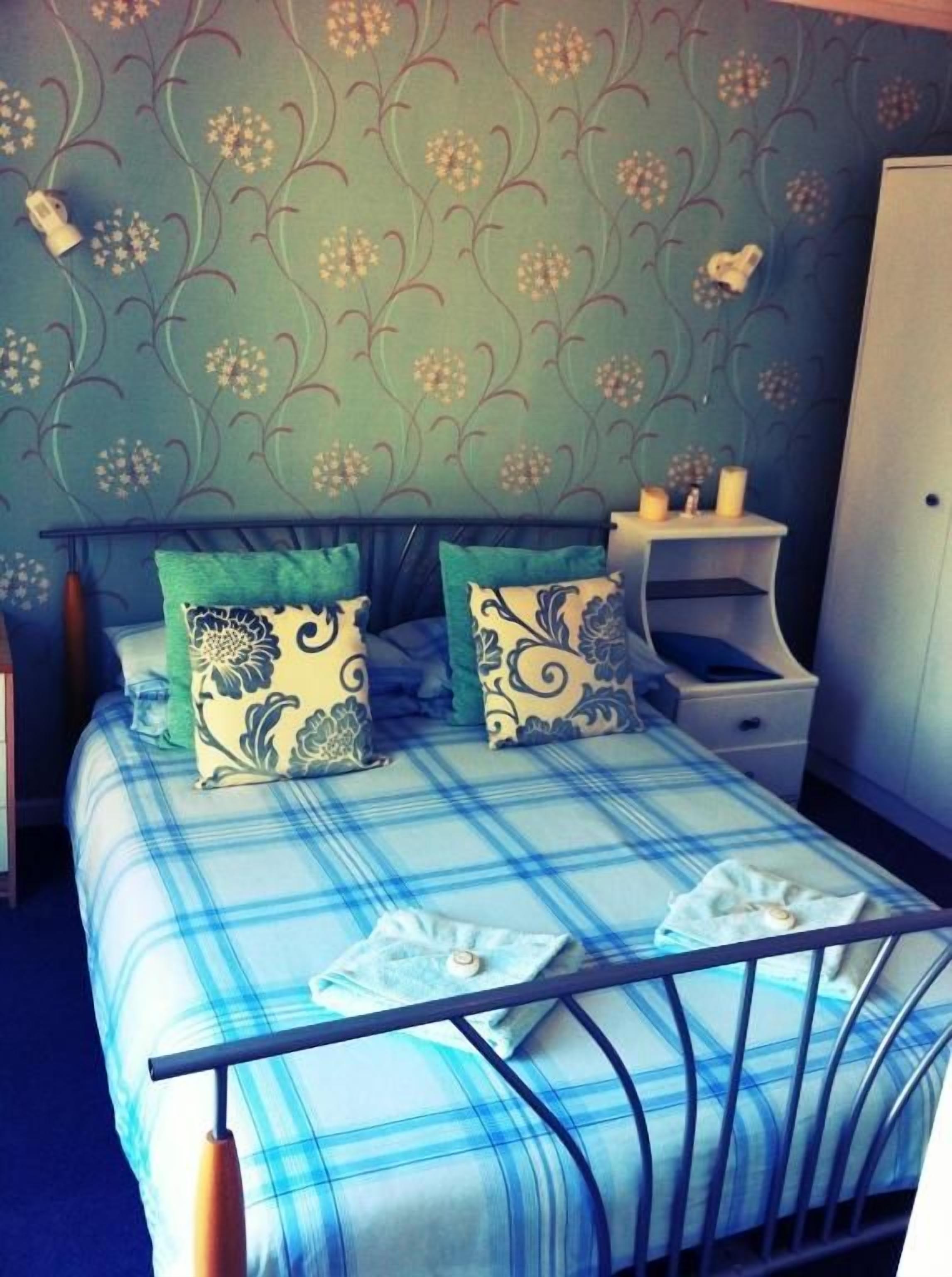 A bed with a blue and white checkered bedspread, two decorative pillows, a bedside table with a lamp, and a wall with a floral pattern.