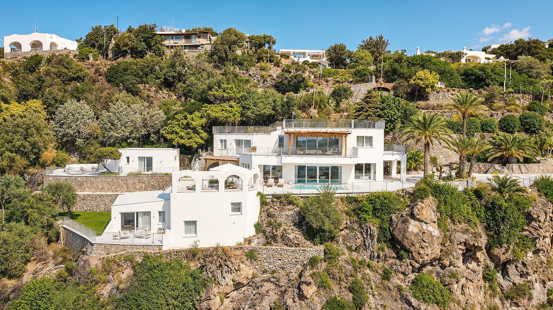 Une maison moderne à plusieurs étages, dotée d’une piscine, entourée d’un terrain rocheux et de végétation.