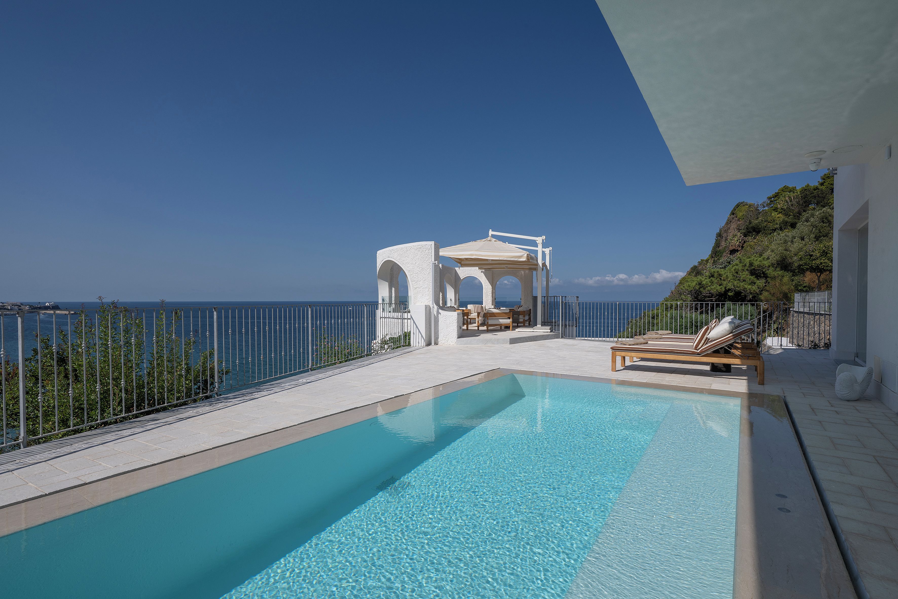 Une piscine d’un bleu éclatant, avec vue sur la mer, un gazebo blanc et des chaises longues.