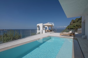 Ein klares, blaues Schwimmbecken mit Blick auf das Meer, ein weißes Pavillon und Liegestühle.