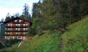 A multi-story wooden building with balconies, surrounded by dense forest. - MOUNTAINSIDE ZERMATT APARTMENT WITH STUNNING VALLEY VIEWS (Zermatt)