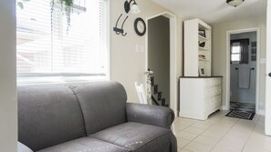 Een woonkamer met een grijze bank, een wit kastje, een klok, en een trap die naar een badkamer leidt.