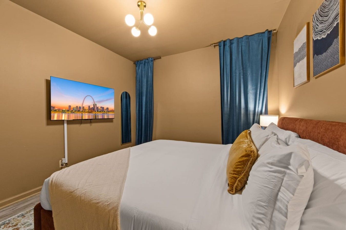 A hotel room with a bed, a flat-screen TV mounted on the wall, a ceiling light fixture, and a window with blue curtains.