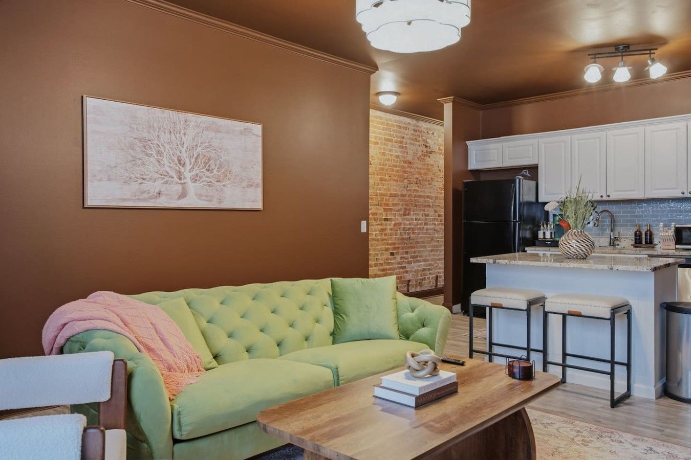 Una sala de estar con un sofá verde, una mesa de centro de madera y una zona de cocina con armarios blancos y un refrigerador negro.