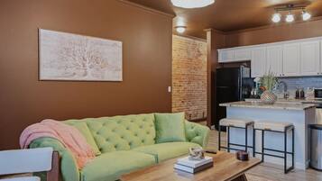 A living room with a green sofa, a wooden coffee table, and a kitchen area with white cabinets and a black refrigerator.