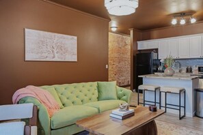 A living room with a green sofa, a wooden coffee table, and a kitchen area with white cabinets and a black refrigerator.