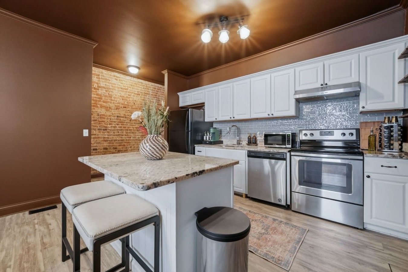 A kitchen with white cabinets, stainless steel appliances, a brick accent wall, and a small bar area with stools.