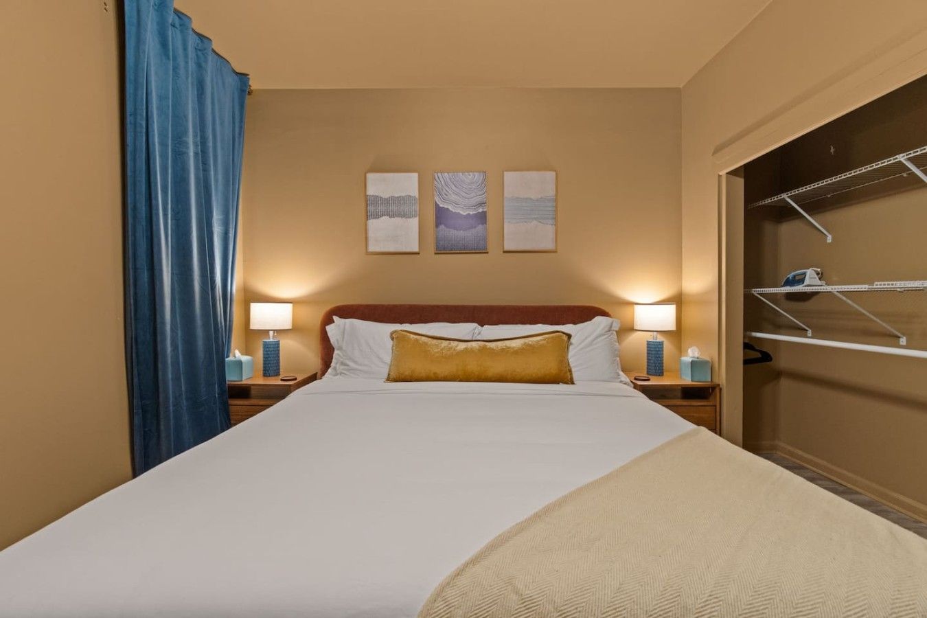 Una habitación de hotel con una cama, dos lámparas de noche, una cortina azul y un armario con una percha.