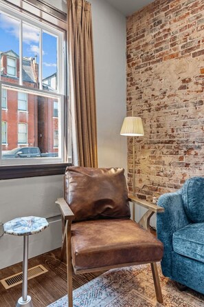 Ein Zimmer mit Ledersessel, blauem Sofa, Lampe, kleinem Tisch und Blick ins Freie durch ein fensterbdecktes Fenster.
