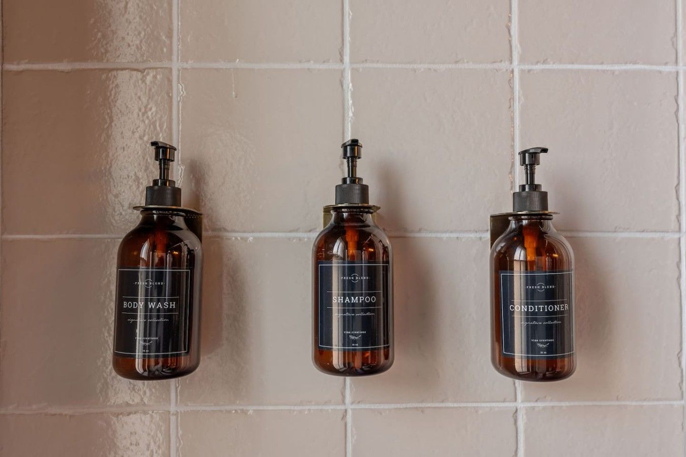 Tre flaconi di bagnoschiuma, shampoo e balsamo su un muro rivestito di piastrelle.