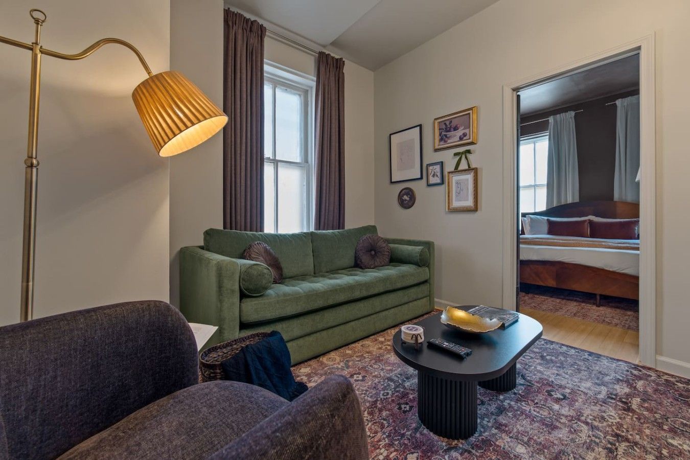 A living room with a green sofa, a coffee table, a floor lamp, and a bed in the adjacent room.