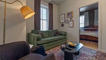 A living room with a green sofa, a coffee table, a floor lamp, and a bed in the adjacent room.