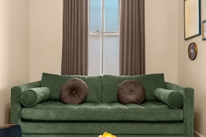 A green sofa with two round cushions in a room with beige walls, a window with curtains, and a framed picture on the wall. - Soulard Gem• 2 min to Farmers Market• 10 min to DT (St. Louis)