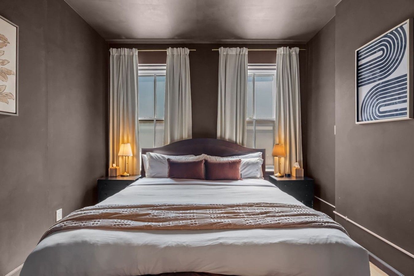 A hotel room with a large bed, two bedside lamps, a view through the window, and a framed abstract artwork on the wall.