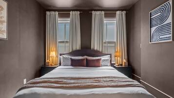 A hotel room with a large bed, two bedside lamps, a view through the window, and a framed abstract artwork on the wall.