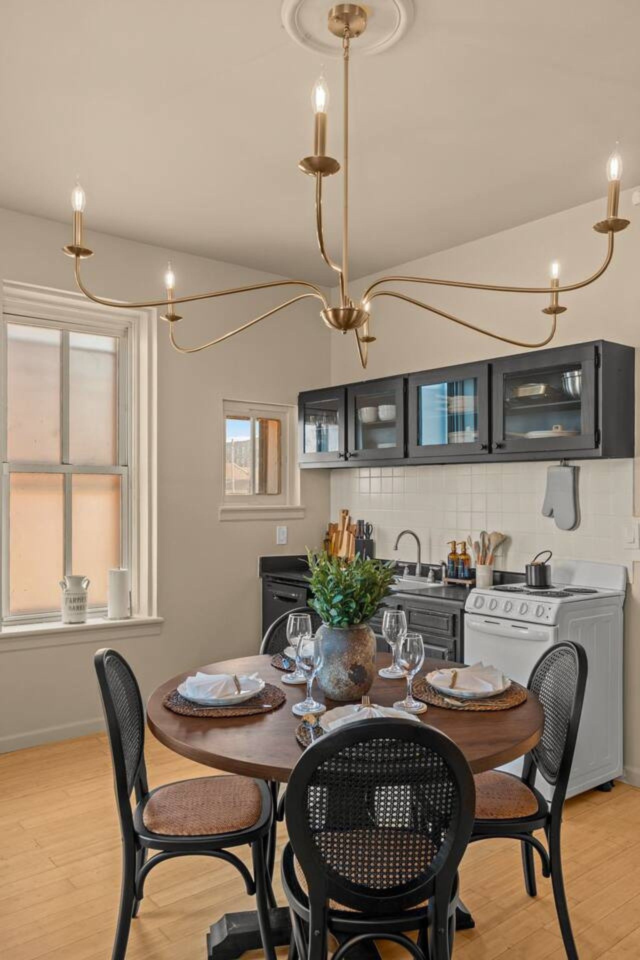 A dining area with a round table, four chairs, a chandelier, and a kitchen in the background.