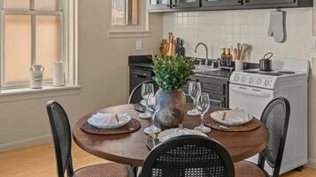 A dining area with a round table, four chairs, a chandelier, and a kitchen in the background.