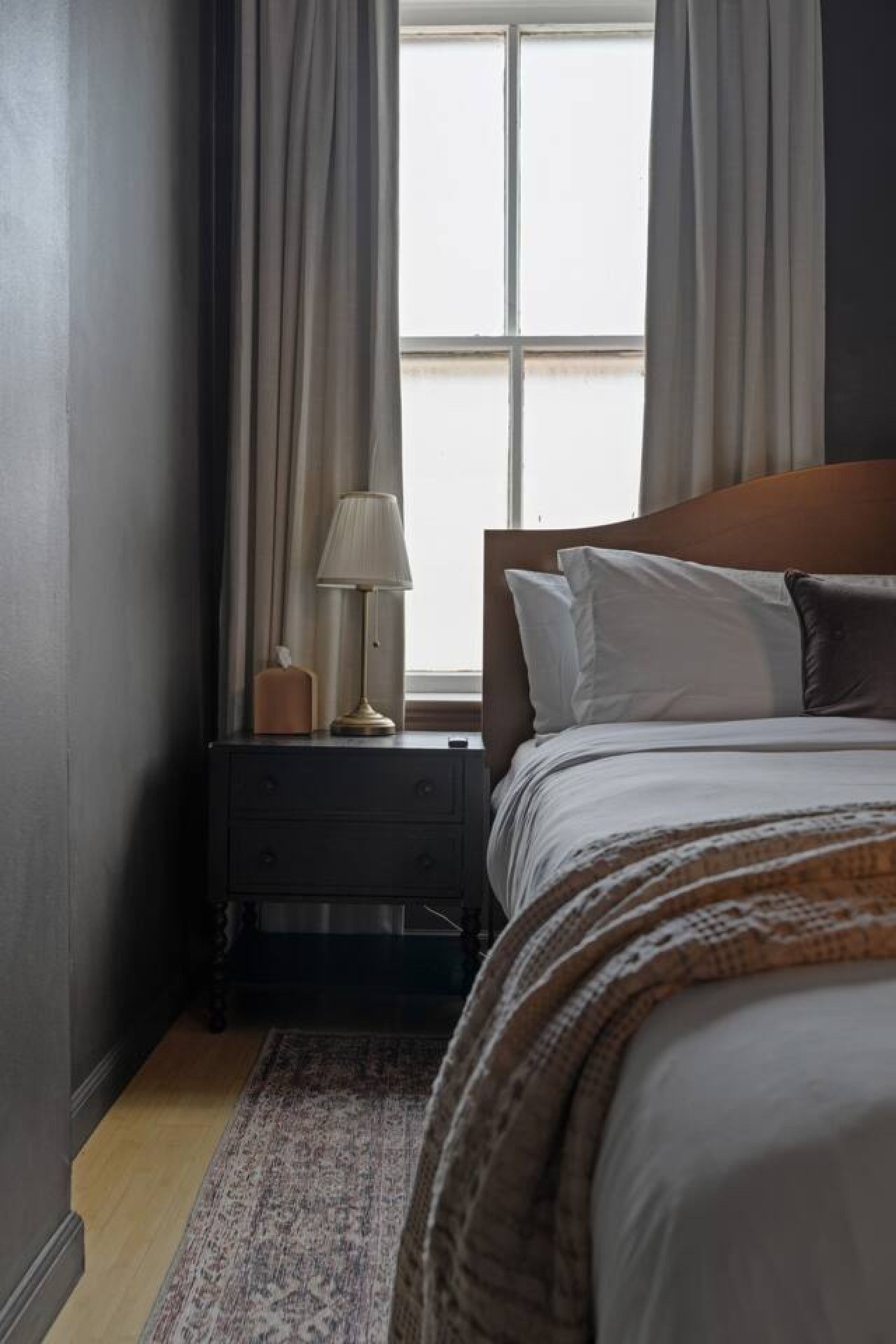 Un dormitorio con una cama, una mesita de noche, una lámpara y una ventana con cortinas.