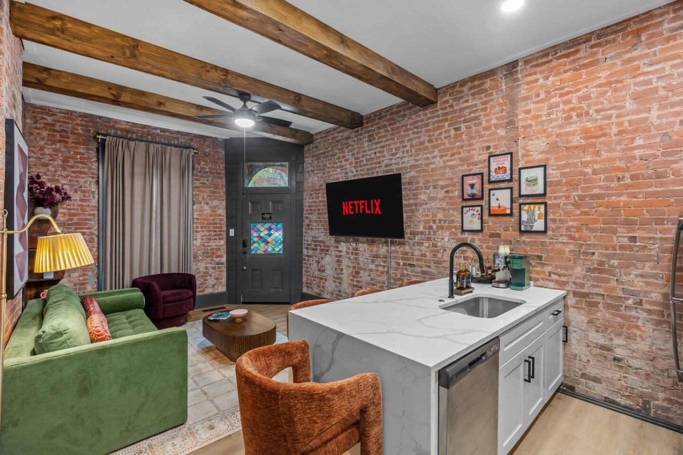 A compact living space with a kitchenette, a green sofa, a red armchair, a ceiling fan, and a door with a colorful window.