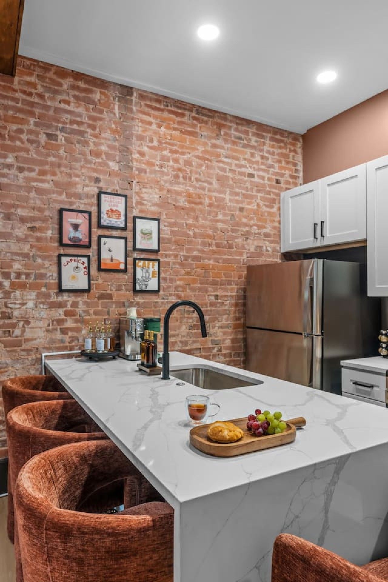 A kitchen with a brick wall, white cabinets, a stainless steel refrigerator, and a marble countertop.
