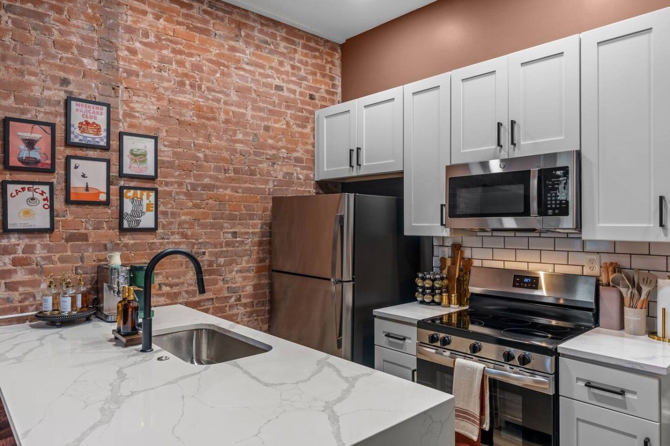 A kitchen with white cabinets, a stainless steel refrigerator, a microwave, and a stove. There are framed pictures on the brick wall.