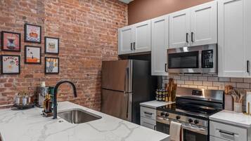 A kitchen with white cabinets, a stainless steel refrigerator, a microwave, and a stove. There are framed pictures on the brick wall.