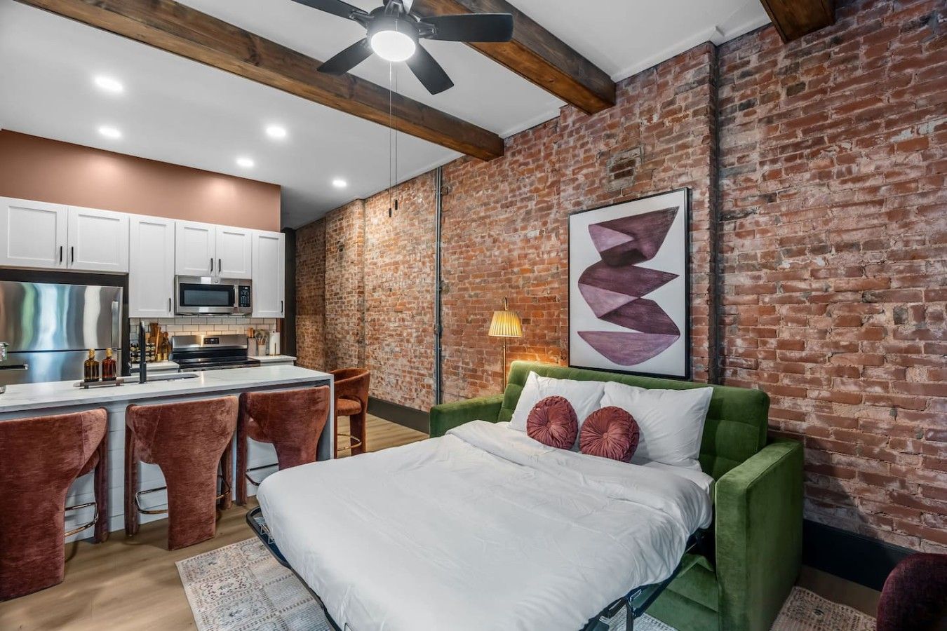 A modern bedroom with a green sofa bed, a large bed, and a kitchen area with white cabinets and a brick wall.