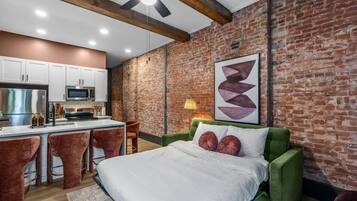 A modern bedroom with a green sofa bed, a large bed, and a kitchen area with white cabinets and a brick wall.