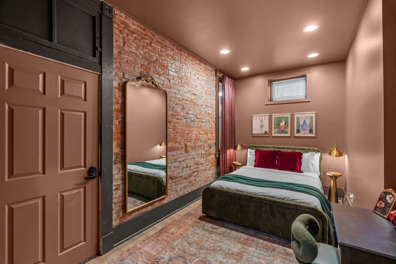 Un dormitorio con pared de ladrillo, cama con ropa de cama verde y roja, escritorio con lámpara y espejo.